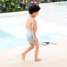 Load image into Gallery viewer, Child in swim shorts standing by a pool with flowers in the background