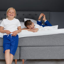 Load image into Gallery viewer, Two children on a gray bed, one sitting and one lying down, both smiling. Both wearing blue Pjama items.
