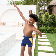 Load image into Gallery viewer, Child wearing blue and yellow swim trunks standing on a stone path outdoors.
